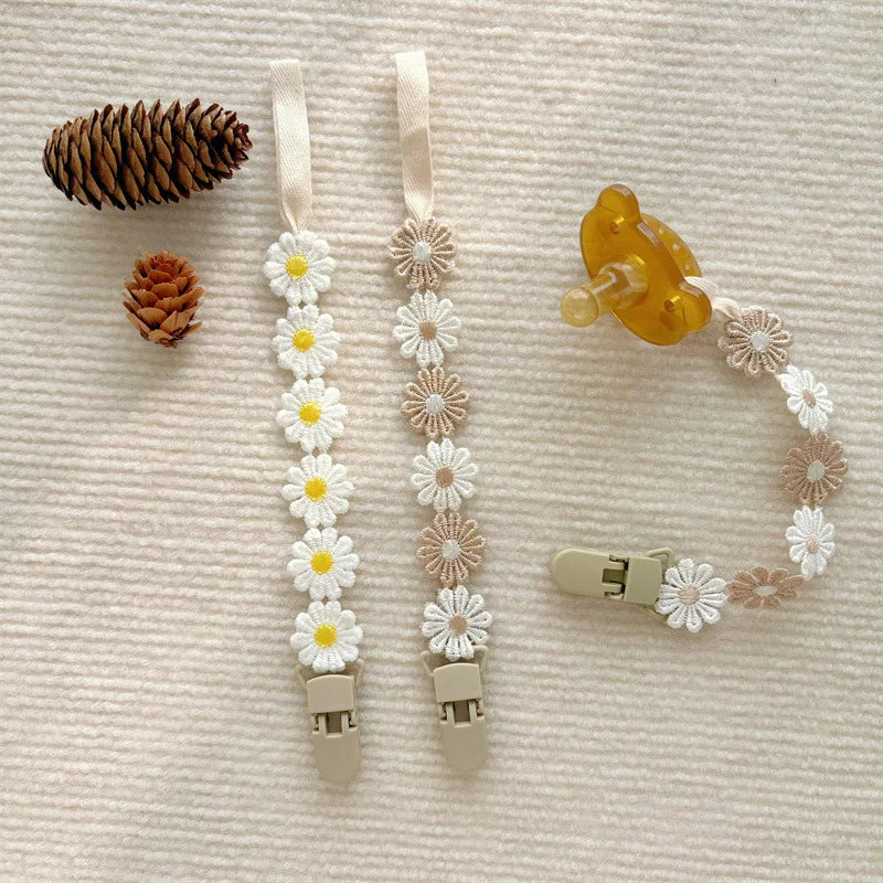 Two daisy pacifier clips, one white and one beige, with a matching pacifier chain and a yellow pacifier.