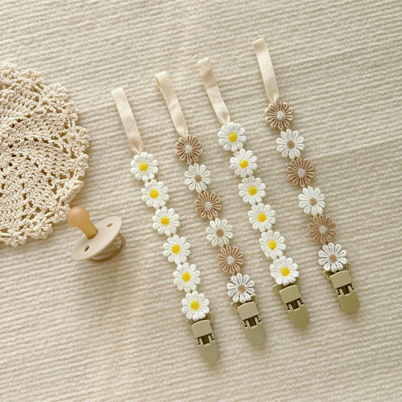 Four daisy flower pacifier clips in white, yellow, and beige, with a pacifier and crochet mat.