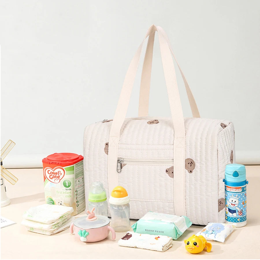 Stylish quilted beige diaper bag with brown bear embroidery, shown with baby bottles, formula, diapers, and toys.