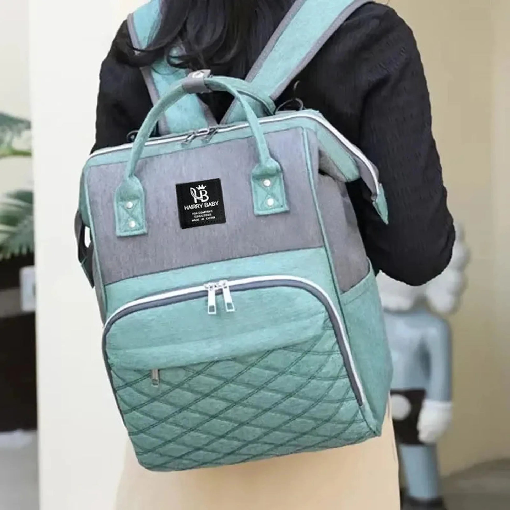 Two-tone gray and teal oxford fabric diaper backpack with quilted front pocket, worn by a person.