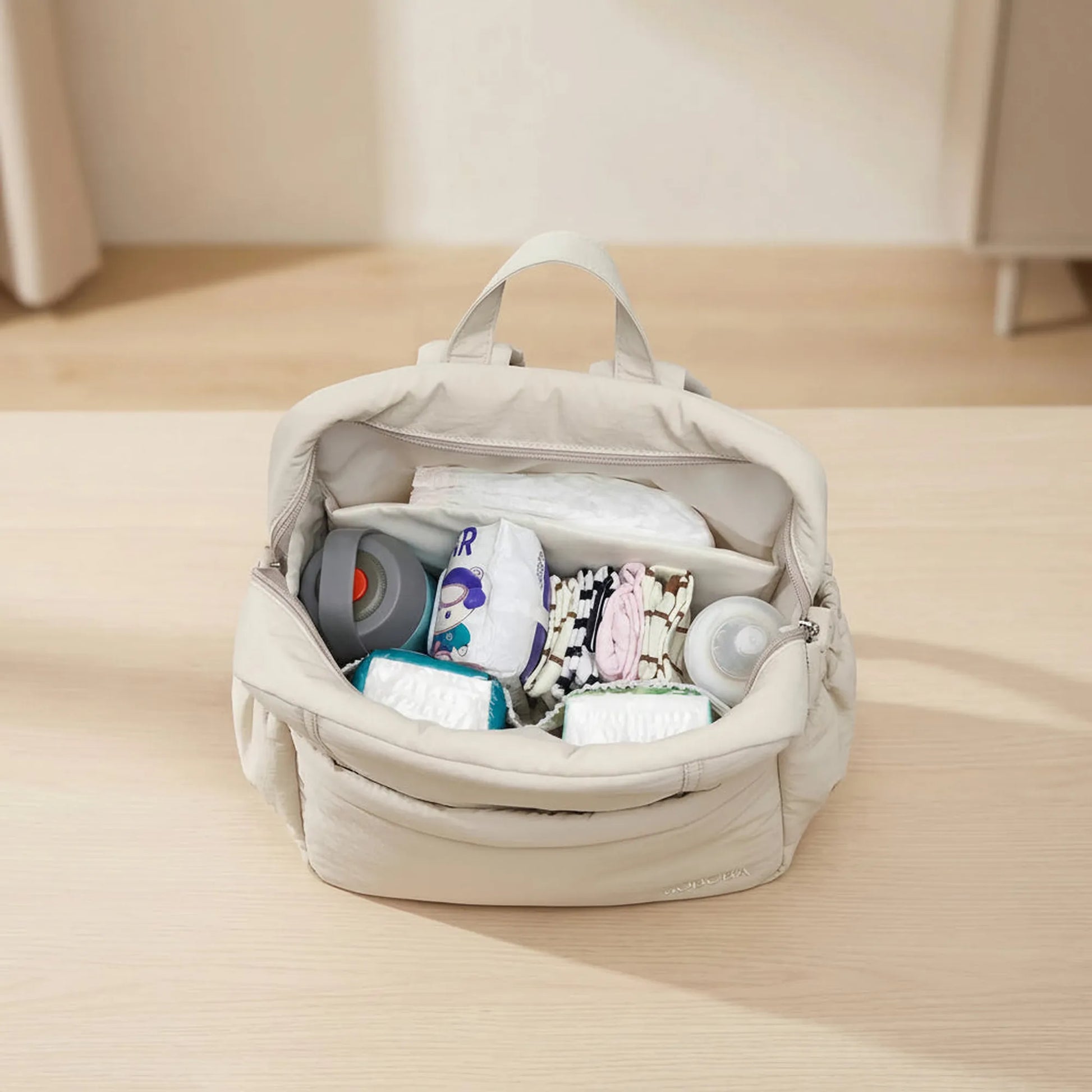 Beige multi-functional diaper backpack, open and filled with baby essentials like diapers, bottles, and clothes.