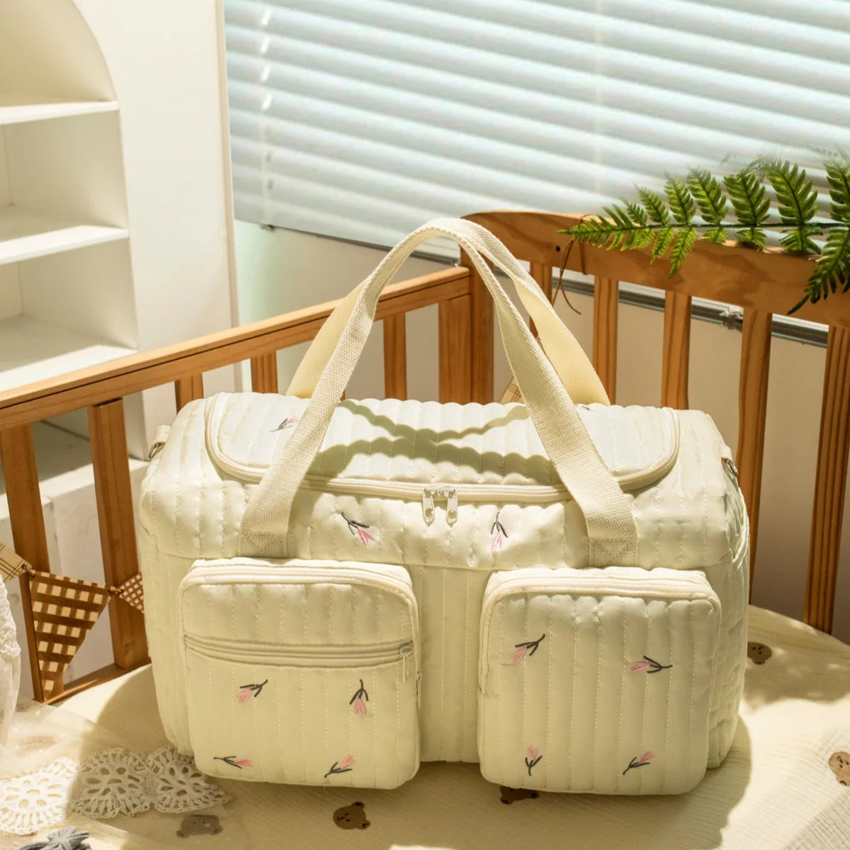 Quilted cream cotton diaper bag with embroidered pink flowers, zipper closure, and front pockets.