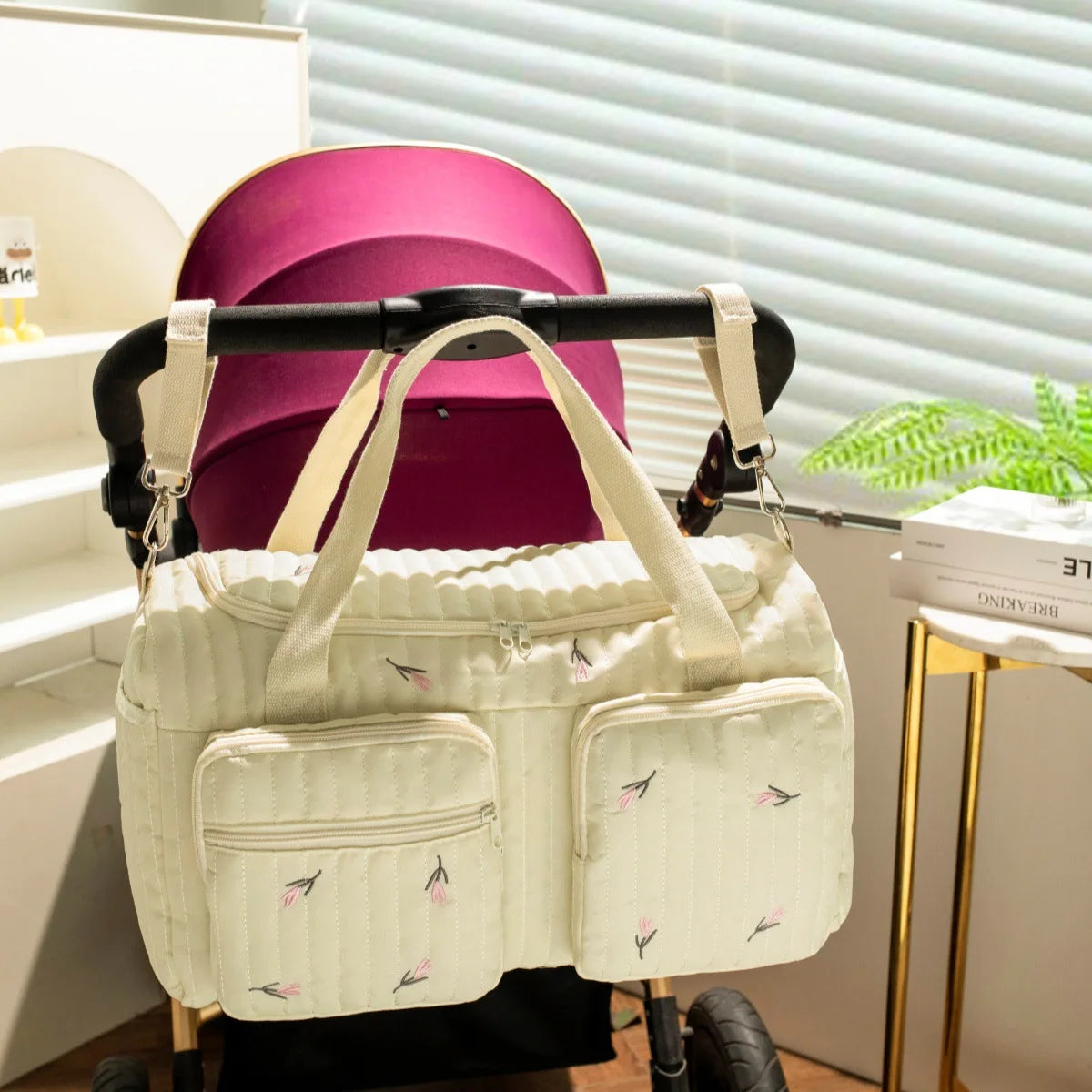 Quilted cream cotton diaper bag with pink floral embroidery, zipper closure, and front pockets, hanging on a stroller.