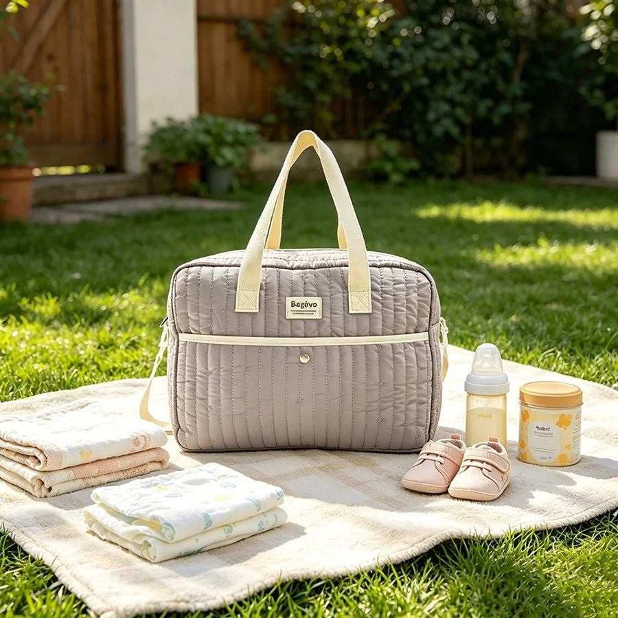 Quilted gray diaper bag with yellow straps, baby shoes, bottle, and formula on a blanket outdoors.