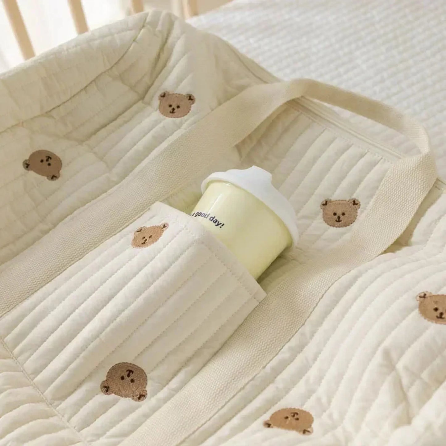 Cream quilted cotton diaper bag with embroidered teddy bears and a bottle in a side pocket.