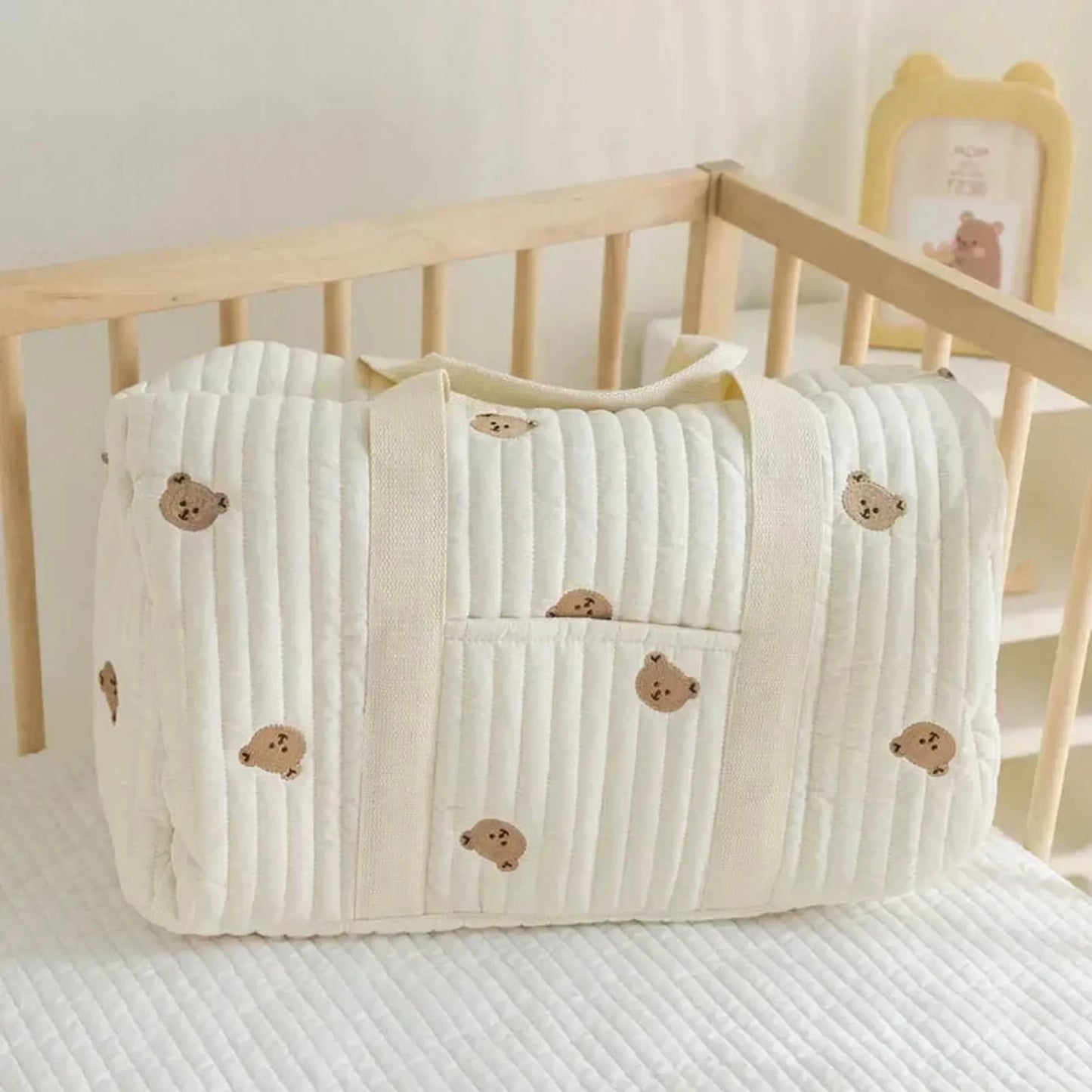 Quilted cream cotton diaper bag with embroidered teddy bears, handles, and front pocket, resting in a crib.