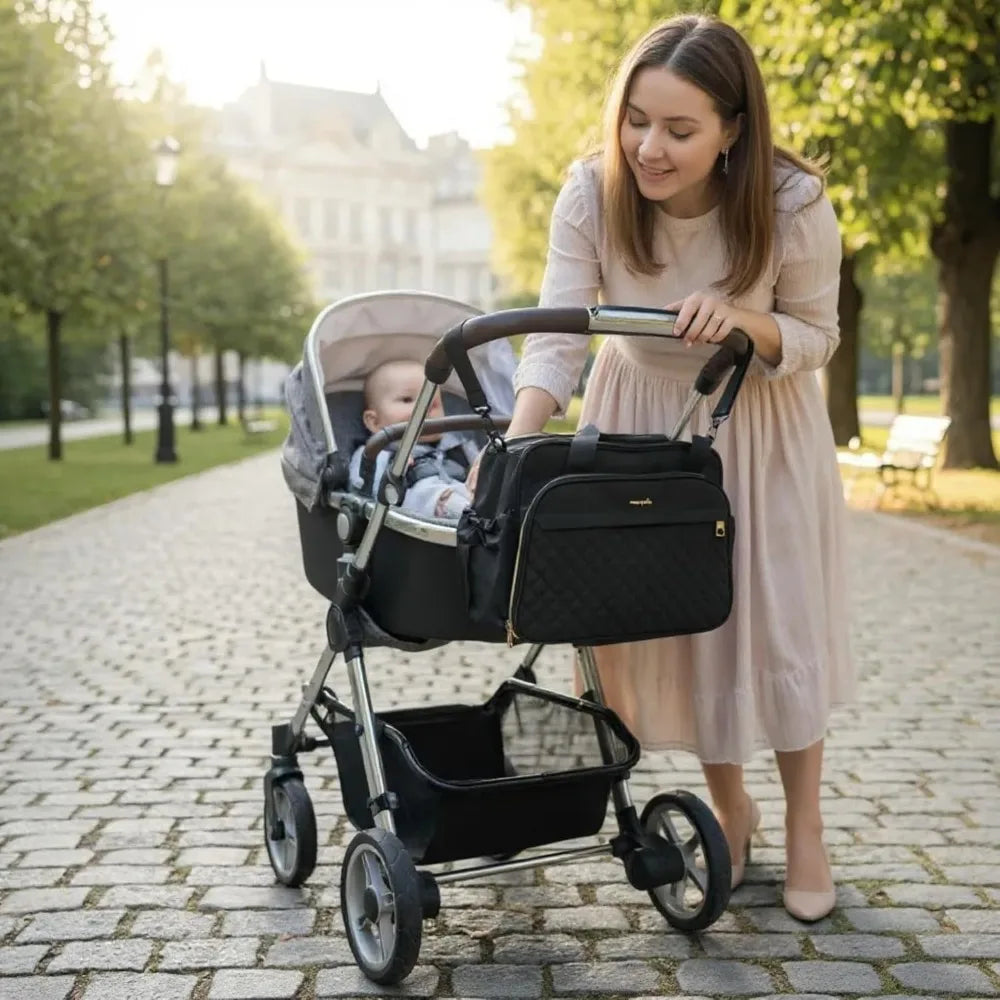 Black quilted diaper bag attached to a stroller, with a mother tending to her baby outdoors.
