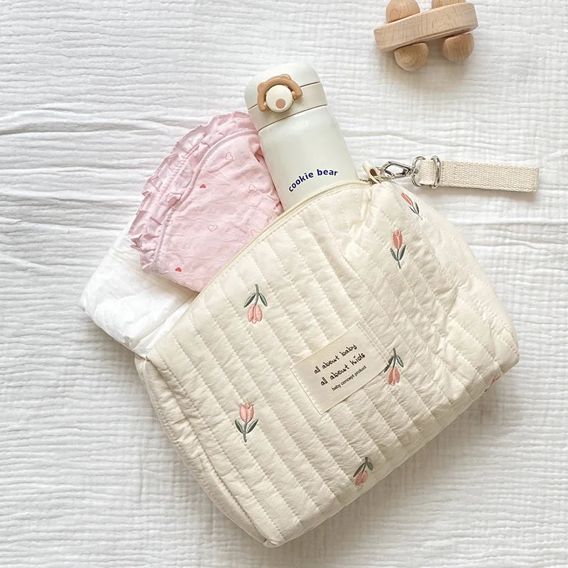 Quilted cream cotton diaper bag with pink tulip embroidery, zipper, and wrist strap, holding a white bottle.